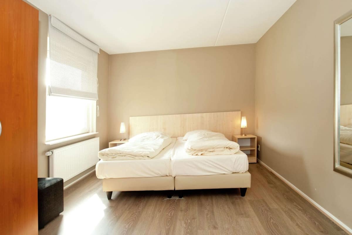 Une chambre d’hôtel avec un lit double, des lampes de chevet, une tête de lit en bois, un radiateur et une fenêtre avec des stores.