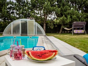 A poolside setting with a glass table holding watermelon slices and drinks, a covered pool, and a hammock in the background. - Comfortable Apartment in Nemojov Near Forest (Nemojov)