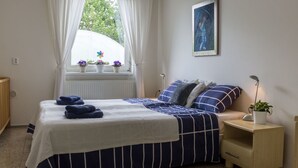 Un dormitorio con cama, mesita de noche, cortinas en la ventana, un radiador y un cuadro en la pared.