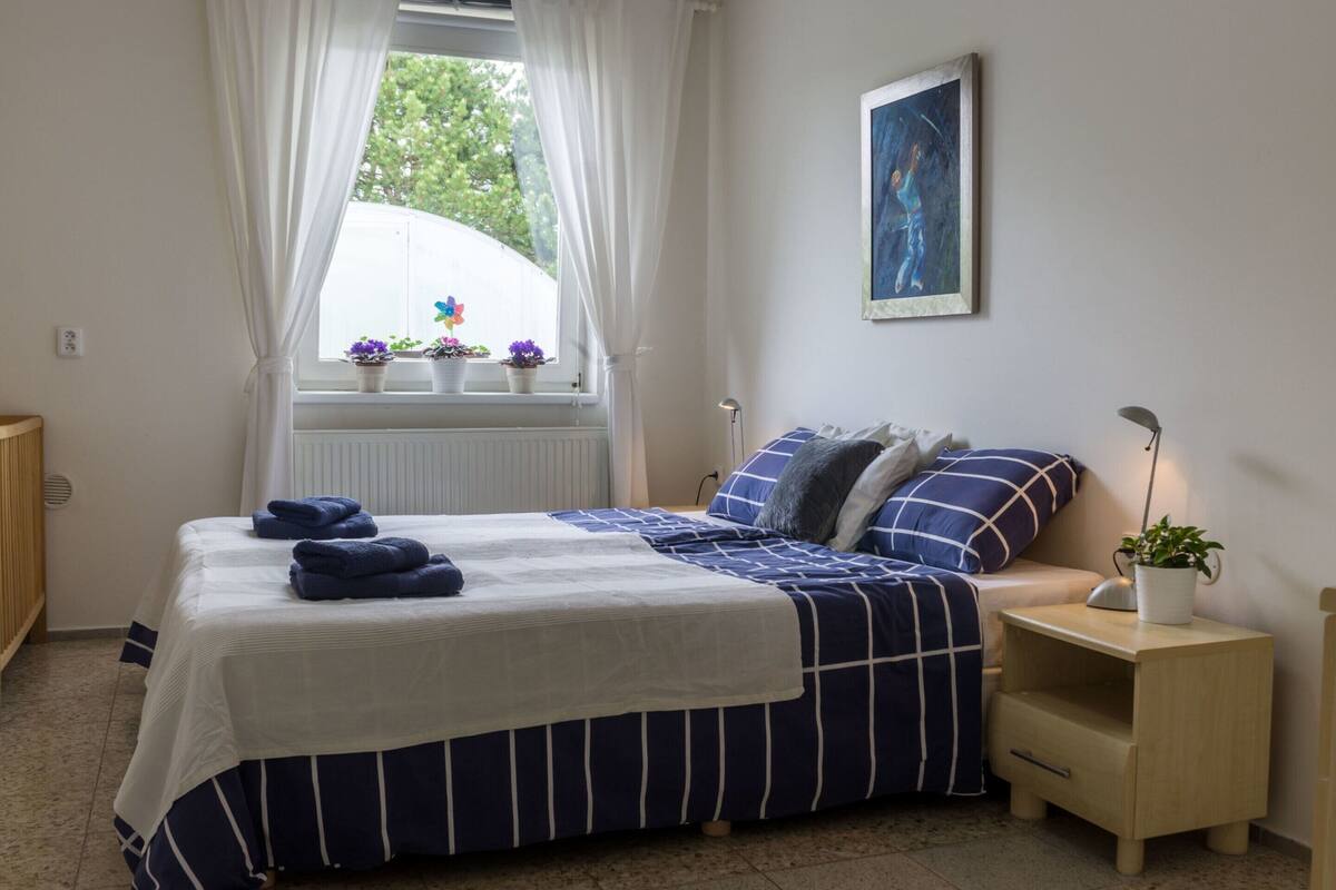 Une chambre à coucher avec un lit, une table de chevet, une commode, une fenêtre avec des rideaux, un radiateur et un tableau au mur.