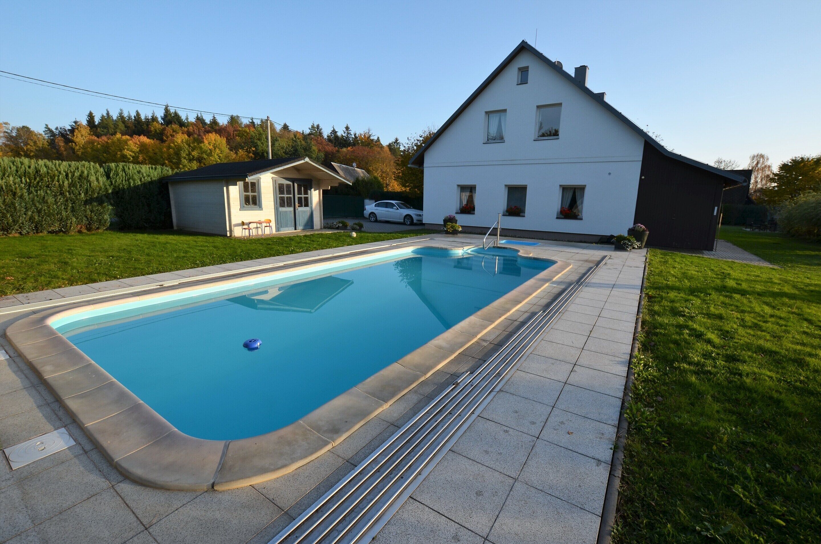 A rectangular swimming pool with a tiled deck, surrounded by a well-maintained lawn and a house in the background.