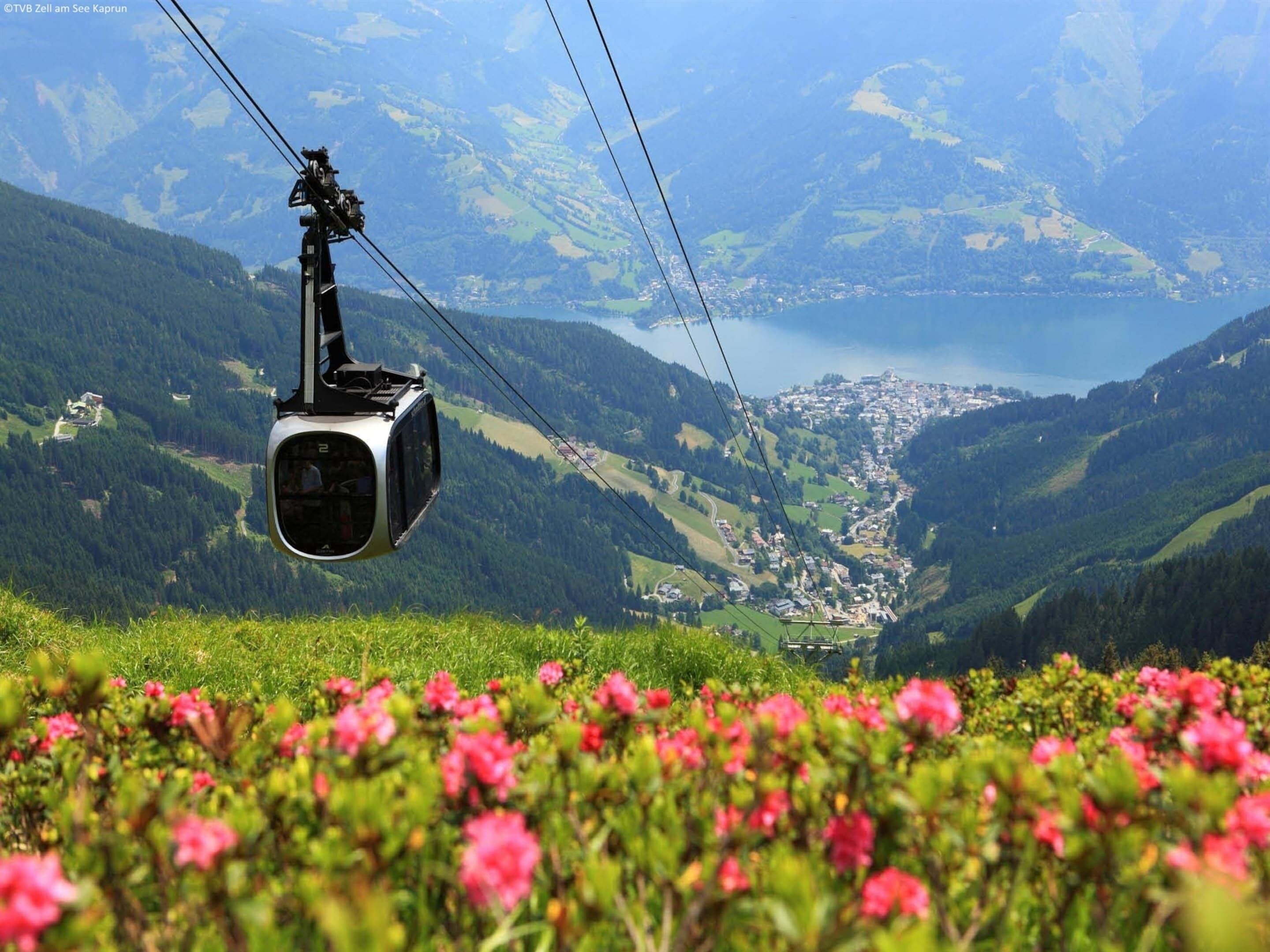 A cable car suspended over a valley with a town below and a lake in the distance.