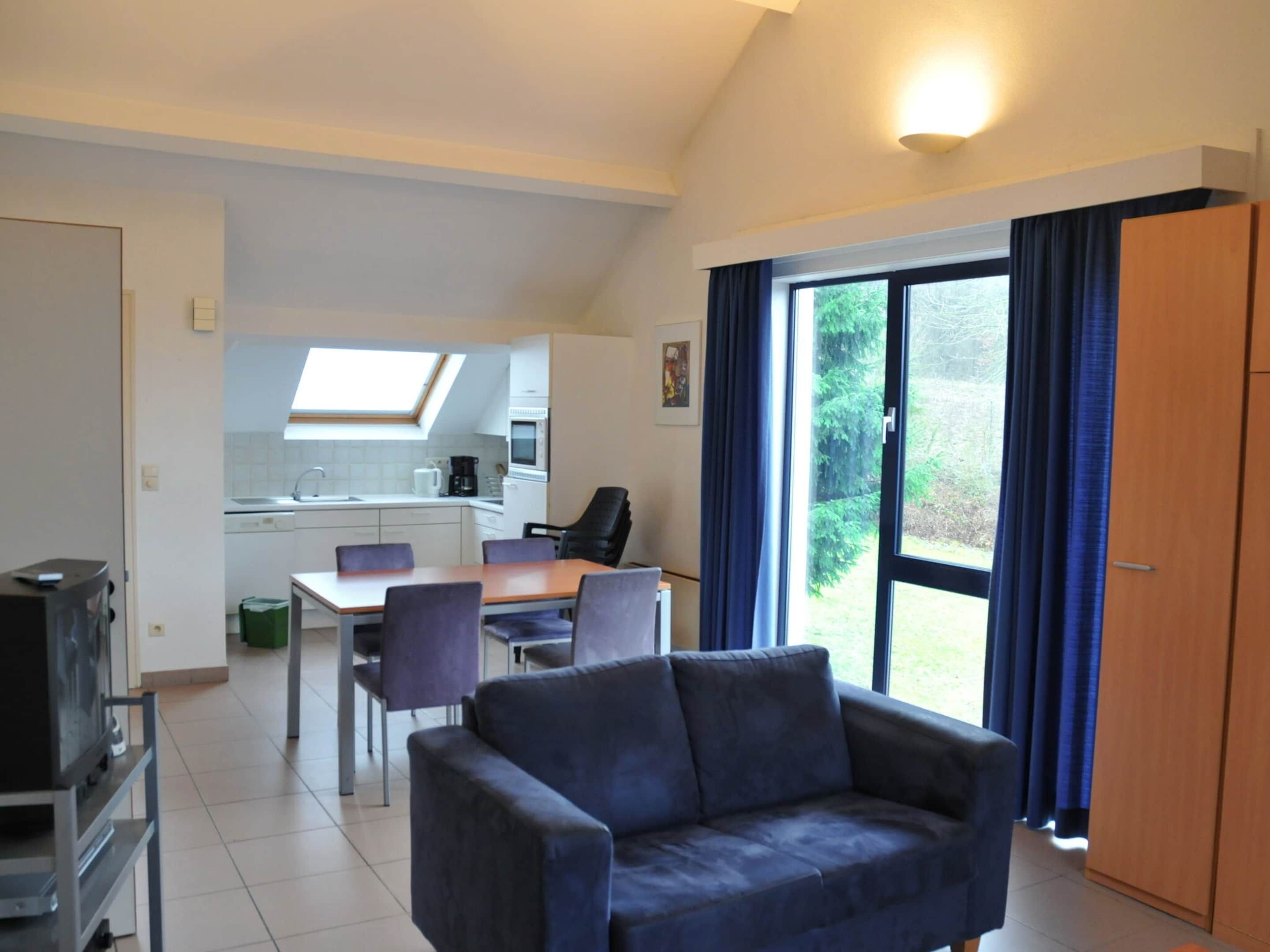 A modern living room with a sofa, a dining table with chairs, a kitchen in the background, and a large window with curtains.