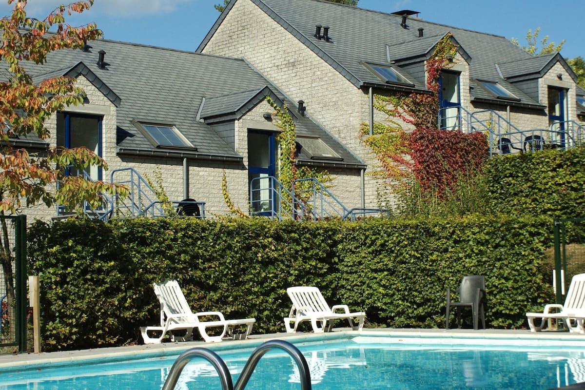 Ett poolområde med vita solstolar och en byggnad i sten med flera fönster och ett sluttande tak.