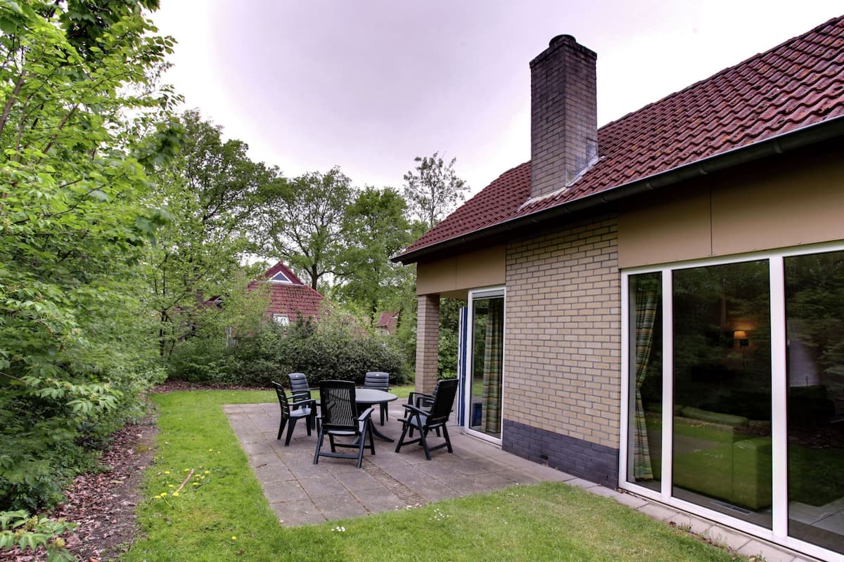 Eine Terrasse mit Tisch und Stühlen, ein Ziegelkamin und eine Glastür, die zu einer grünen Rasenfläche führt.