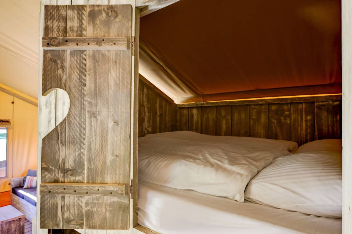 Ein schlichtes hölzernes Bett, ein kleines Holzregal und Blick in einen gemütlichen Raum mit Sofa und Fenster.