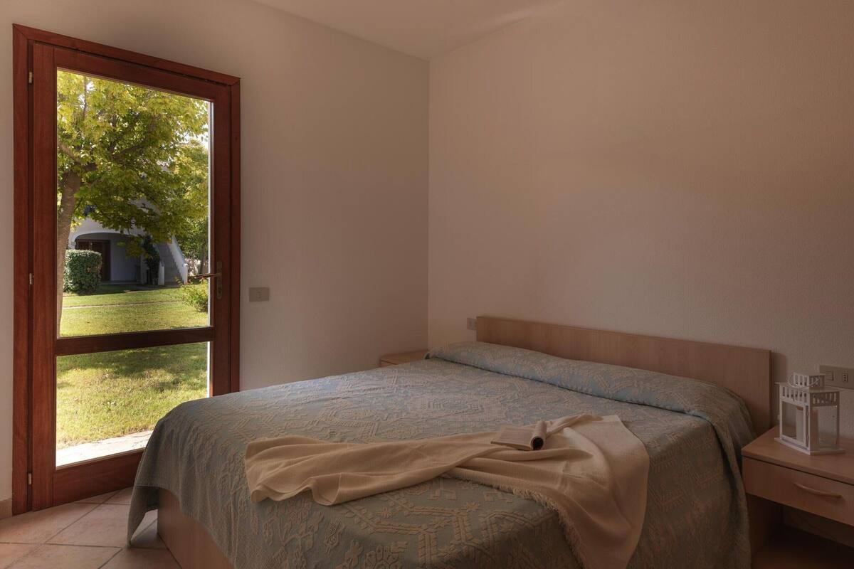 A bedroom with a bed, a nightstand, and a window with a view of a tree and a building outside.
