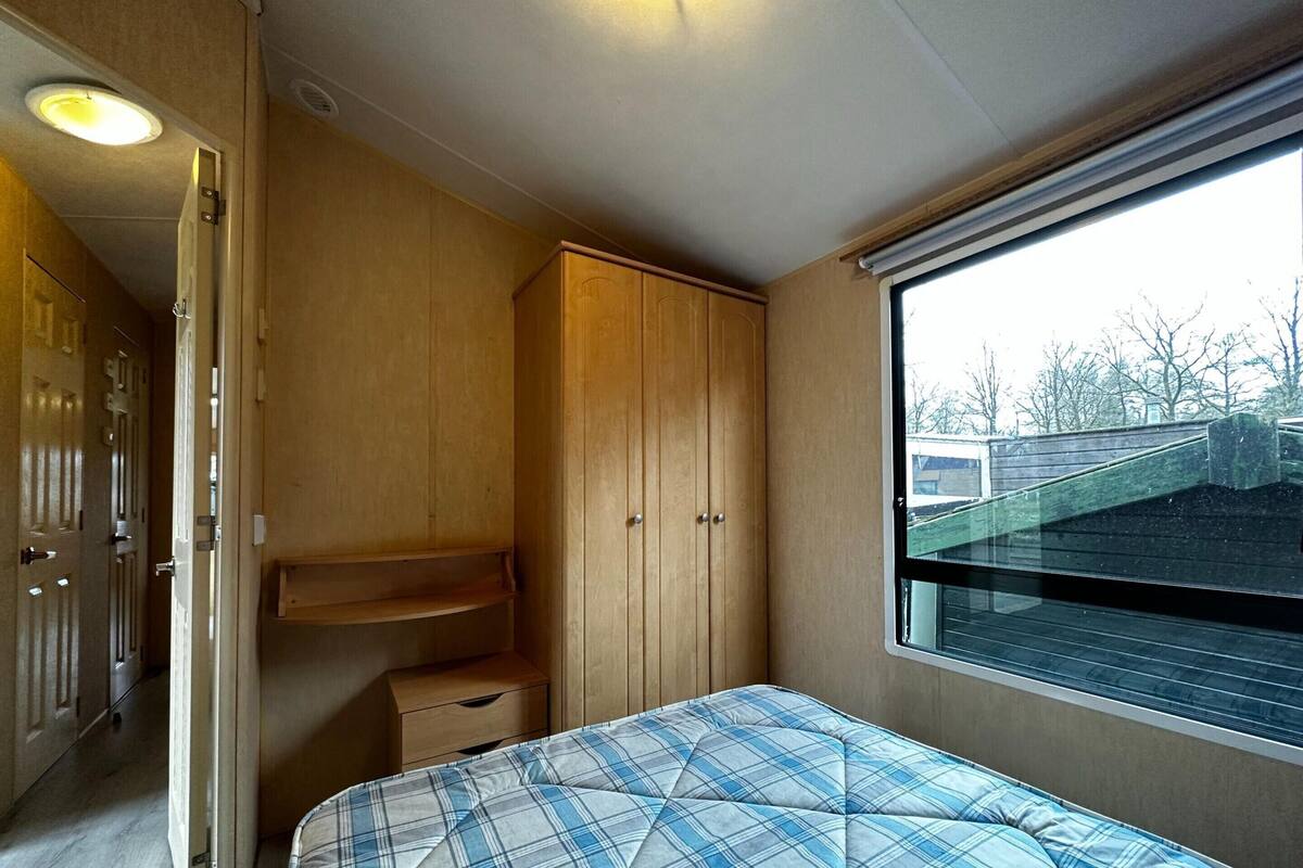 A compact RV interior with a bed, wooden cabinets, and a window providing a view of the outside.