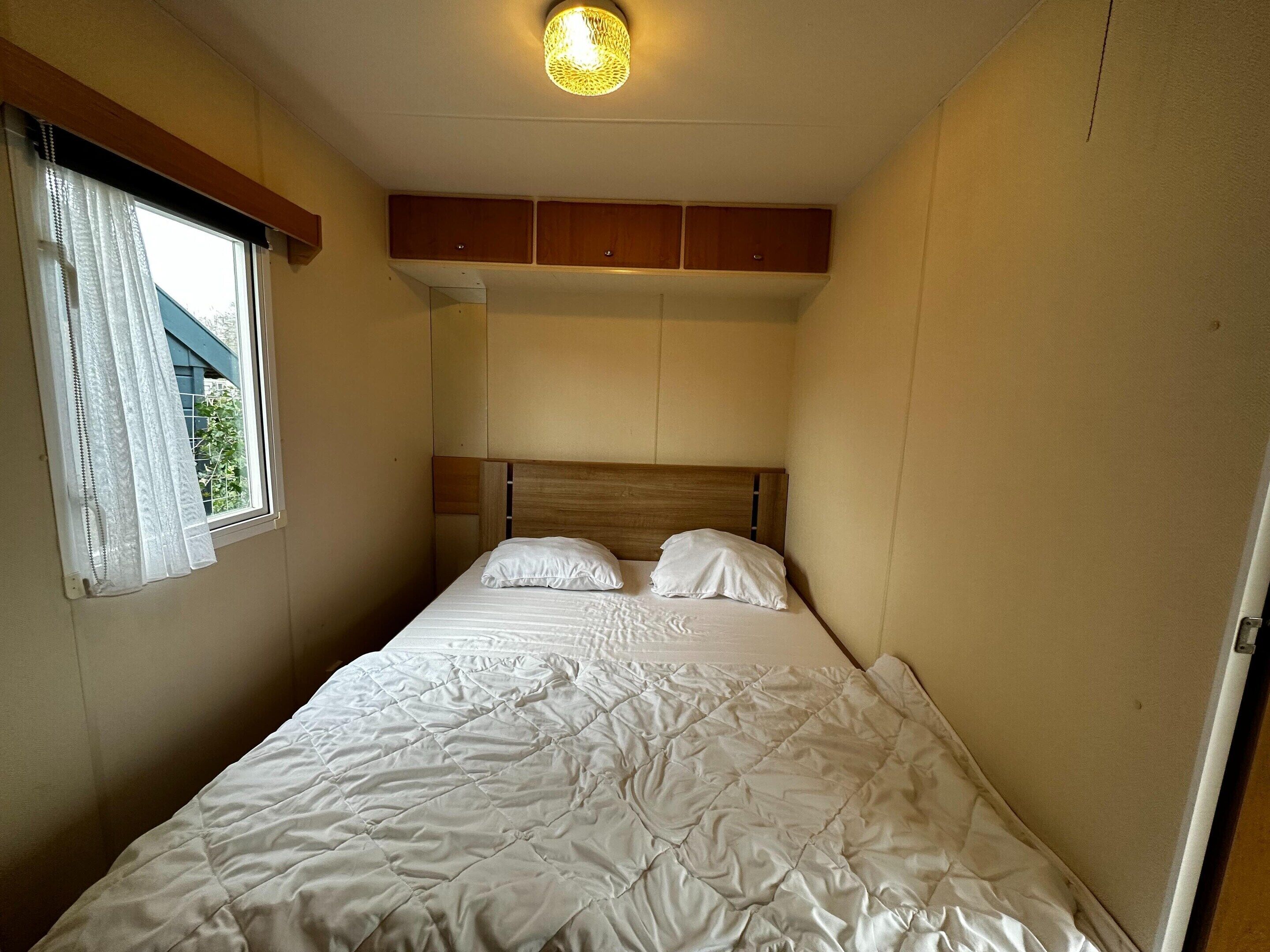 Een enkel bed met een houten hoofdeinde, wit beddengoed en een raam met dunne gordijnen.