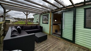 Ein überdachter Außenbereich mit Ecksofa und kleinem Tisch, einer Holzterrasse und einem grünen Gebäude mit einer Schiebetür.