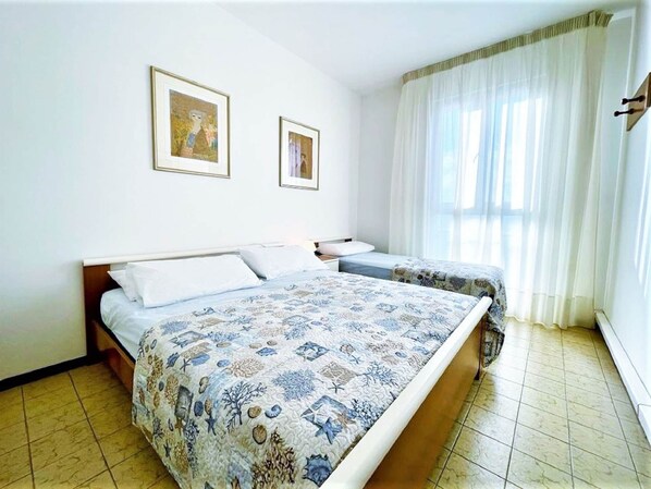 Un dormitorio con dos camas, un ventanal con cortinas translúcidas y dos cuadros en la pared.