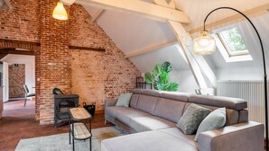 A loft-style room with exposed brick walls, a loft bed, a gray sofa, a wooden bench, and a large window with a view of greenery. - Nice Apartment on Unesco World Heritage Site Wortel-kolonie (Hoogstraten)
