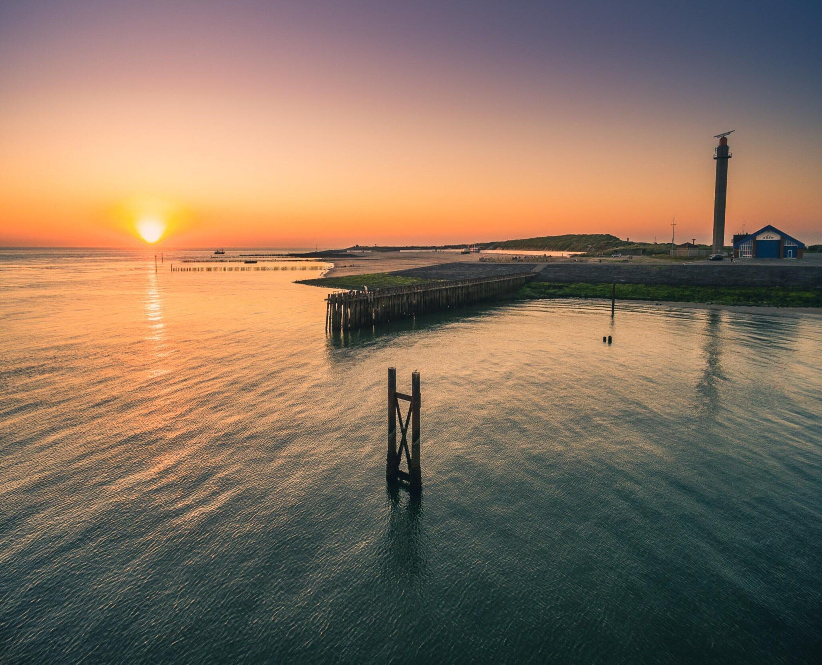 Solnedgang over rolige vand med en ankomstbro, et fyrtårn og en lille bygning ved kysten.