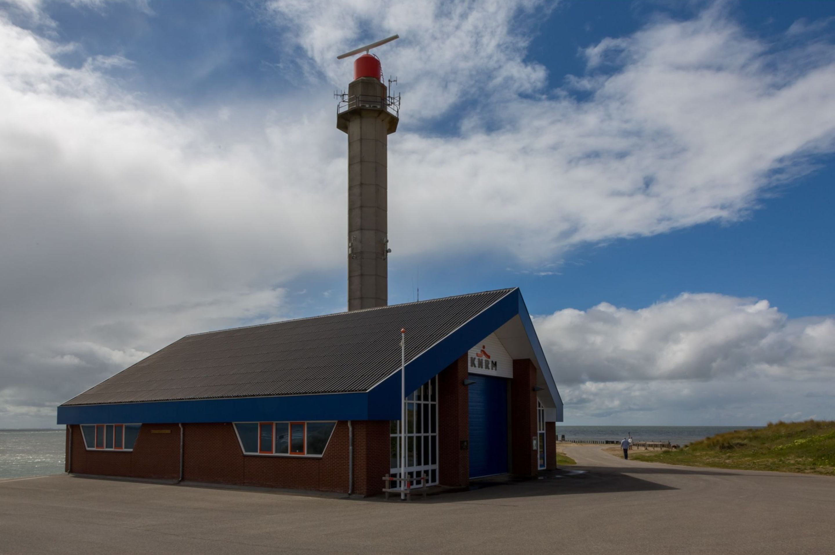A lighthouse with a red top and a blue building with a sloped roof in front.