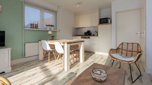 A compact kitchen with white cabinets, a dining area with a wooden table and white chairs, and a wicker chair. - Holiday Home With sun Terrace Facing South-pet-friendly (Westkapelle)
