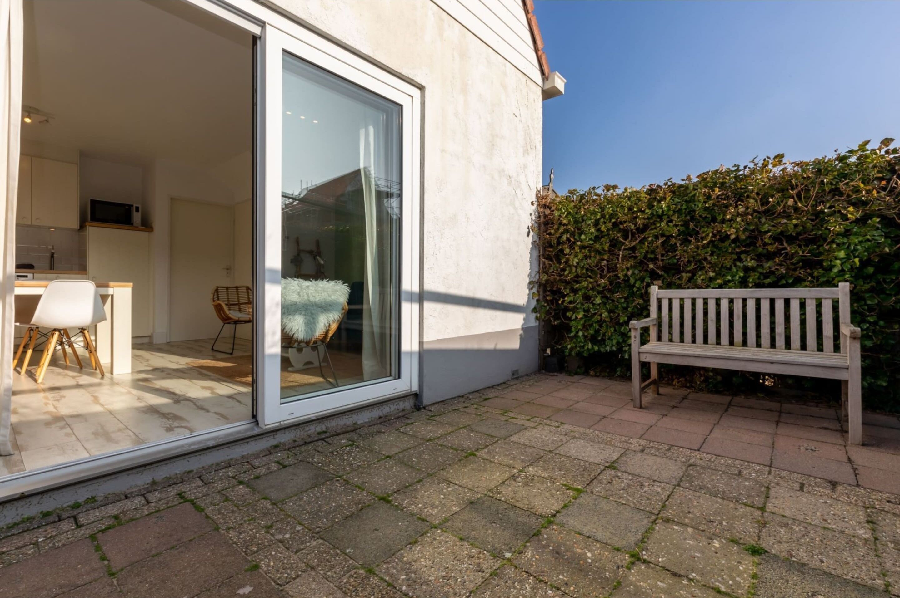 Un patio moderno con puerta de cristal que da a una zona de cocina, un banco de madera y un seto al fondo.