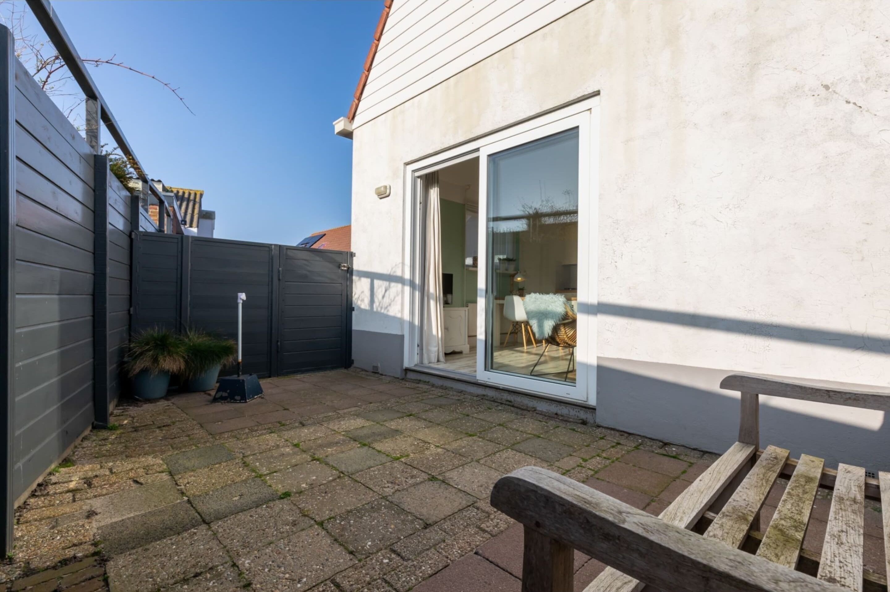 A patio with a wooden bench, a sliding glass door leading to an interior space, and a grey fence.
