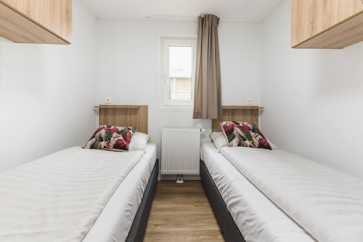 A small, clean room with two single beds, wooden headboards, a window with curtains, and a radiator.