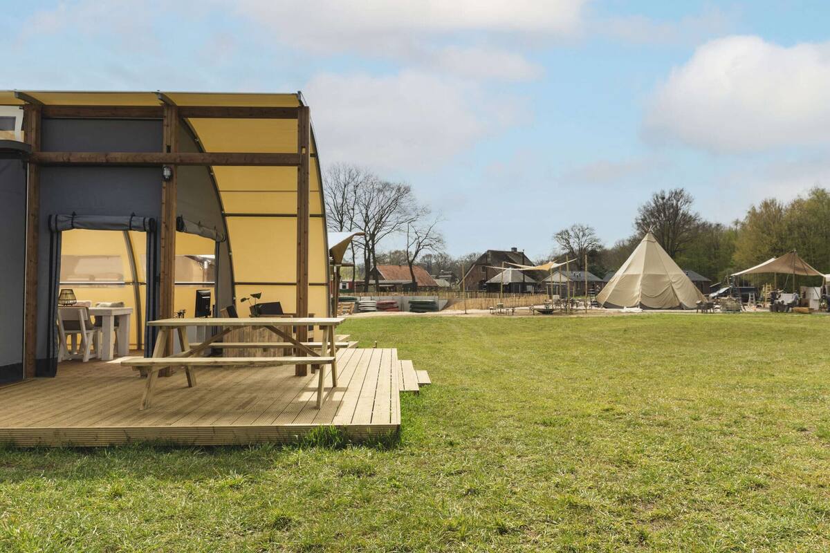A wooden deck with a table and chairs, a large tent, and a teepee in a grassy field with houses in the background.