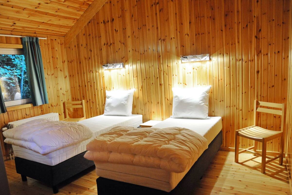 Una habitación de cabaña de madera con dos camas, una silla y un ventanal con cortinas.