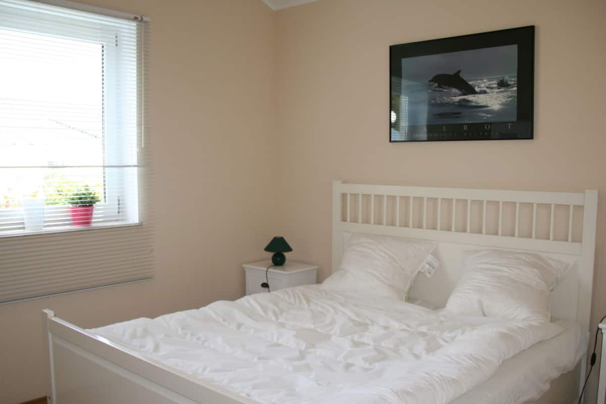 A bedroom with a white bed, a nightstand with a lamp, a window with blinds, and a framed picture on the wall.