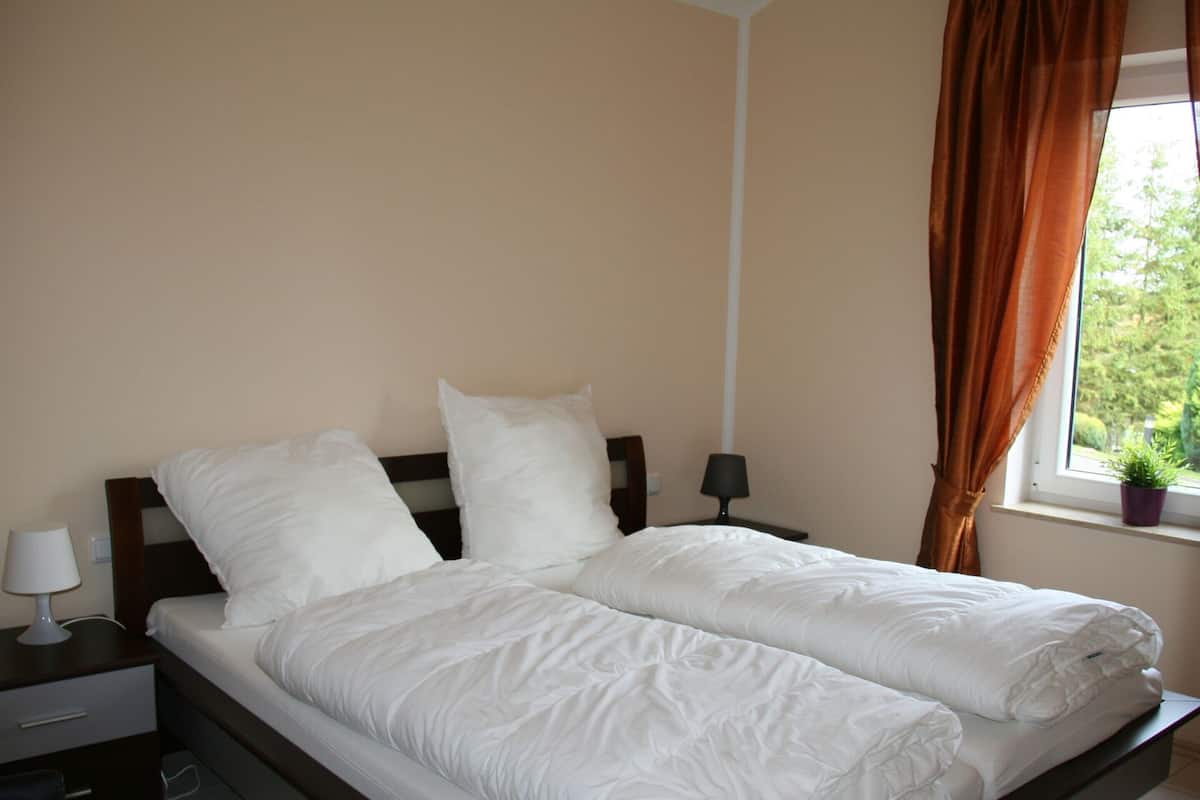 A double bed with white bedding, a nightstand with a lamp, a window with curtains, and a small plant on the windowsill.