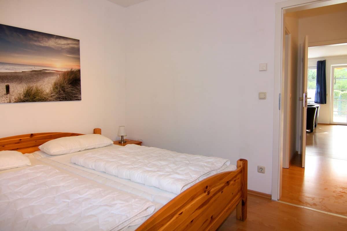 A bedroom with a wooden bed, white bedding, a nightstand with a lamp, and a painting of a beach scene on the wall.