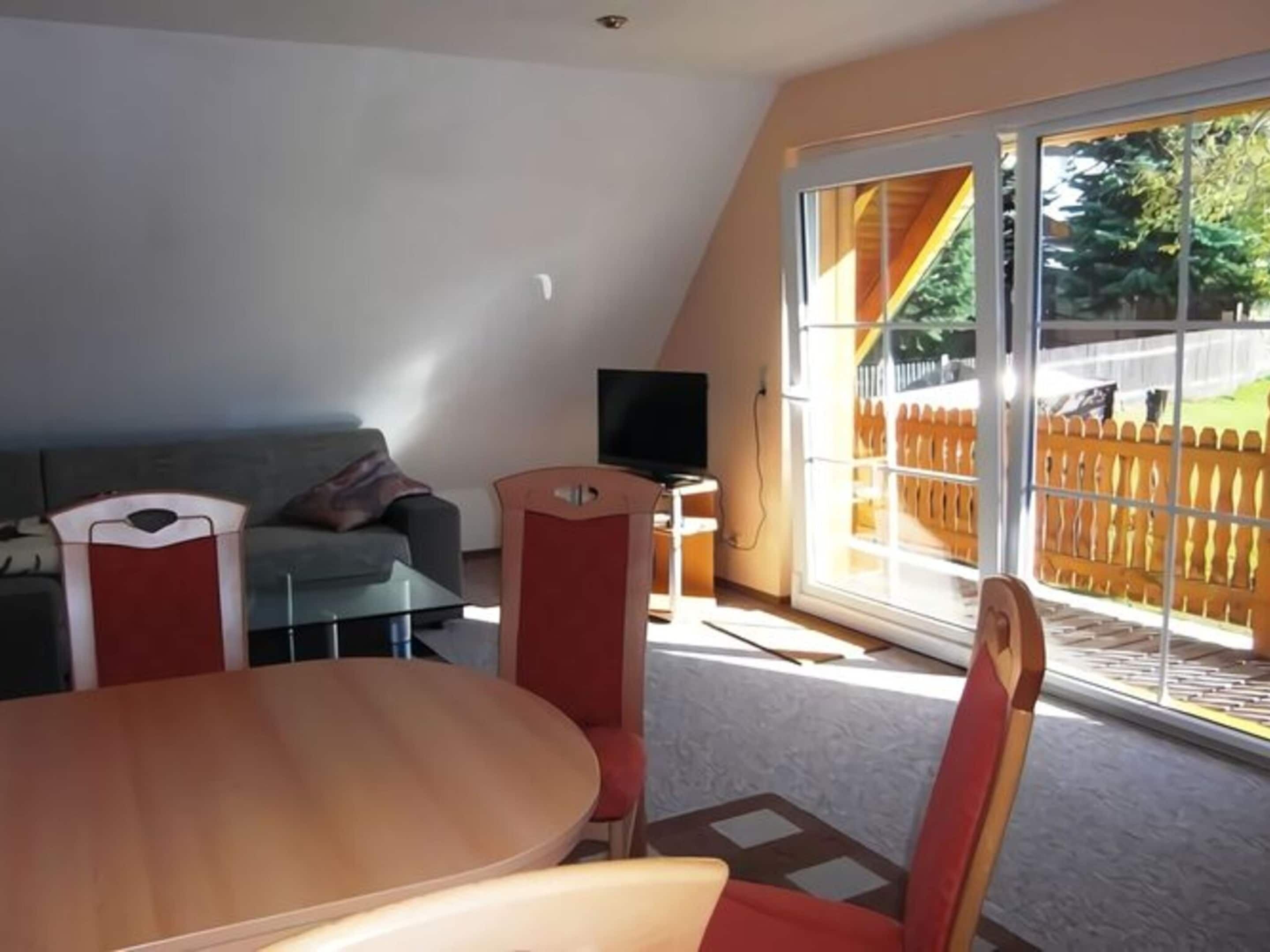 A room with a dining table and chairs, a sofa, a television, and a large window with a view of a wooden fence and greenery.
