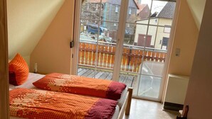 A bedroom with a wooden bed, orange and red bedding, a balcony with a view of houses, and a sliding glass door. - Cozy Apartment in Thale (Thale)