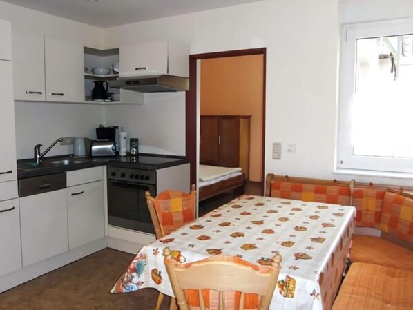 Una cocina compacta con armarios blancos, estufa y pileta. Hay una mesa de comedor con mantel colorido y un banco con cojines anaranjados.