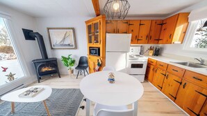 A modern kitchen with wooden cabinets, a white refrigerator, a microwave, and a stove. There is a dining table with chairs, a black and white rug, and a fireplace. - Spa & River view | Le Jardin (Montmagny)