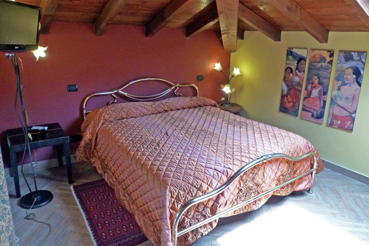 Une chambre à coucher avec un lit, des poutres apparentes en bois au plafond, une table de chevet, une lampe et des œuvres d’art murales.