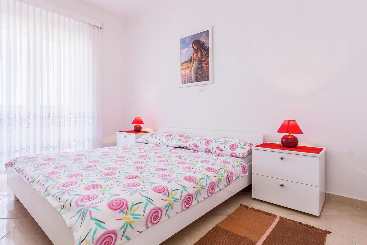 A bedroom with a bed, two nightstands, a red lamp on each, a white wall, and a window with sheer curtains.