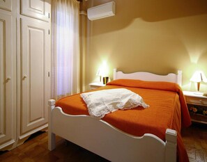 A neatly arranged bedroom with a white bed, orange bedding, and a white lace throw. There are two bedside lamps and a small table with books. - Apartment in San Bartolomeo Al Mare (San Bartolomeo al Mare)