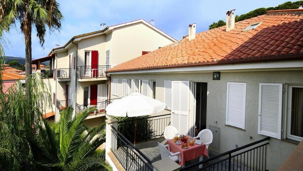 A balcony with a table and chairs, a white umbrella, and a view of neighboring buildings. - Apartment in San Bartolomeo Al Mare (San Bartolomeo al Mare)