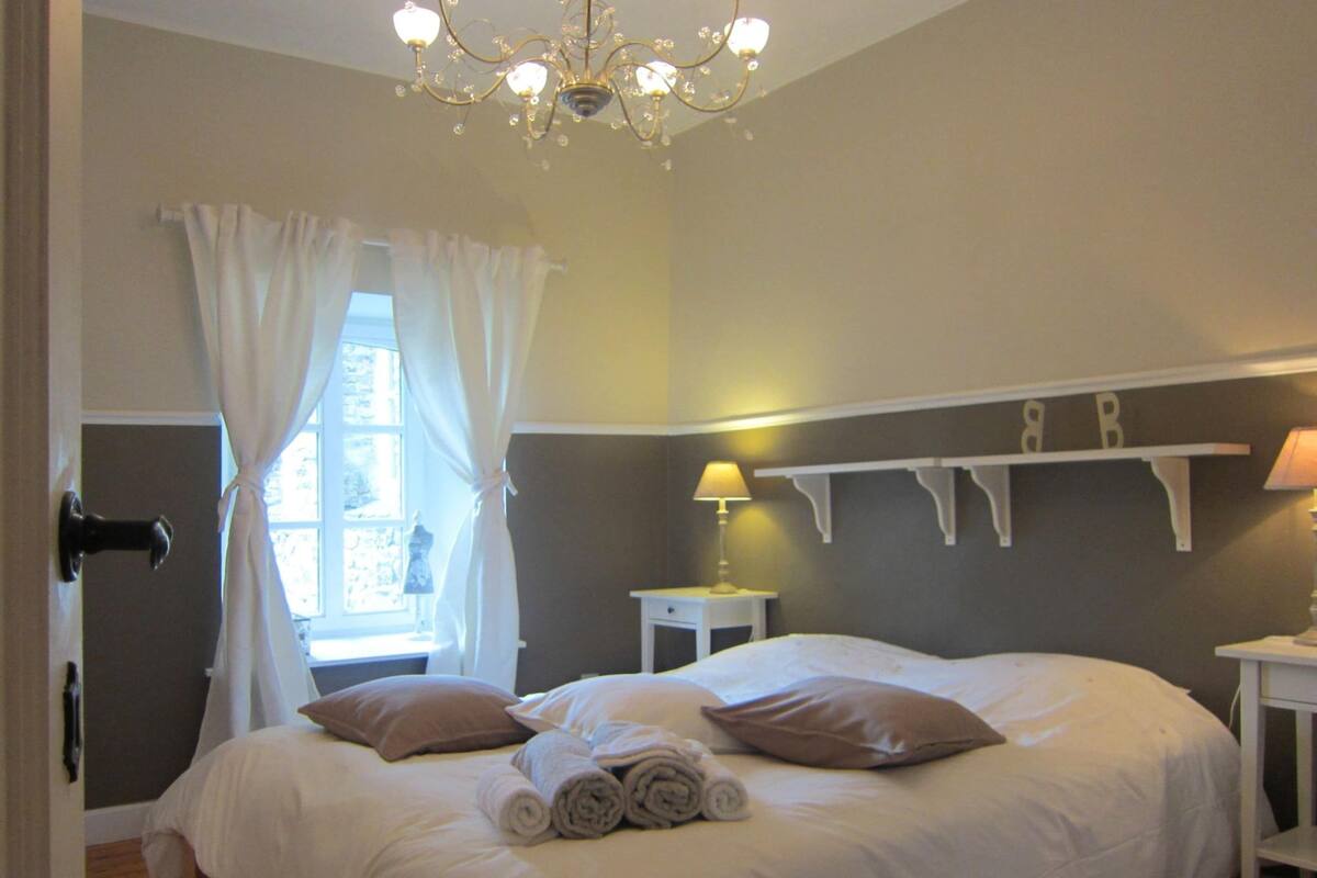 A hotel room with a large bed, a chandelier, a window with curtains, a bedside table with a lamp, and a shelf with decorative items.