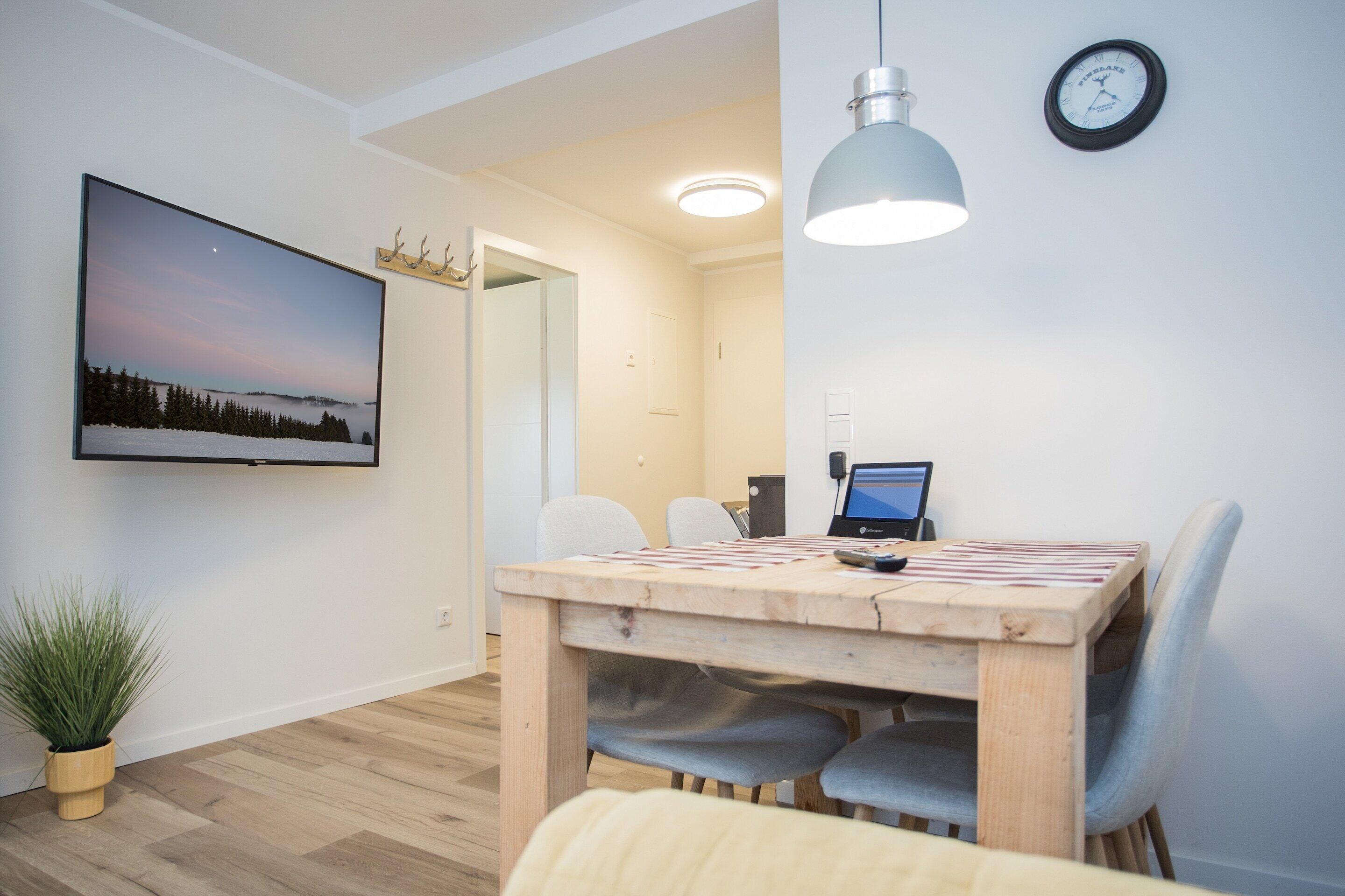 Uma sala de jantar moderna com uma mesa de madeira, cadeiras, uma planta em vaso, uma TV fixada na parede e um relógio.