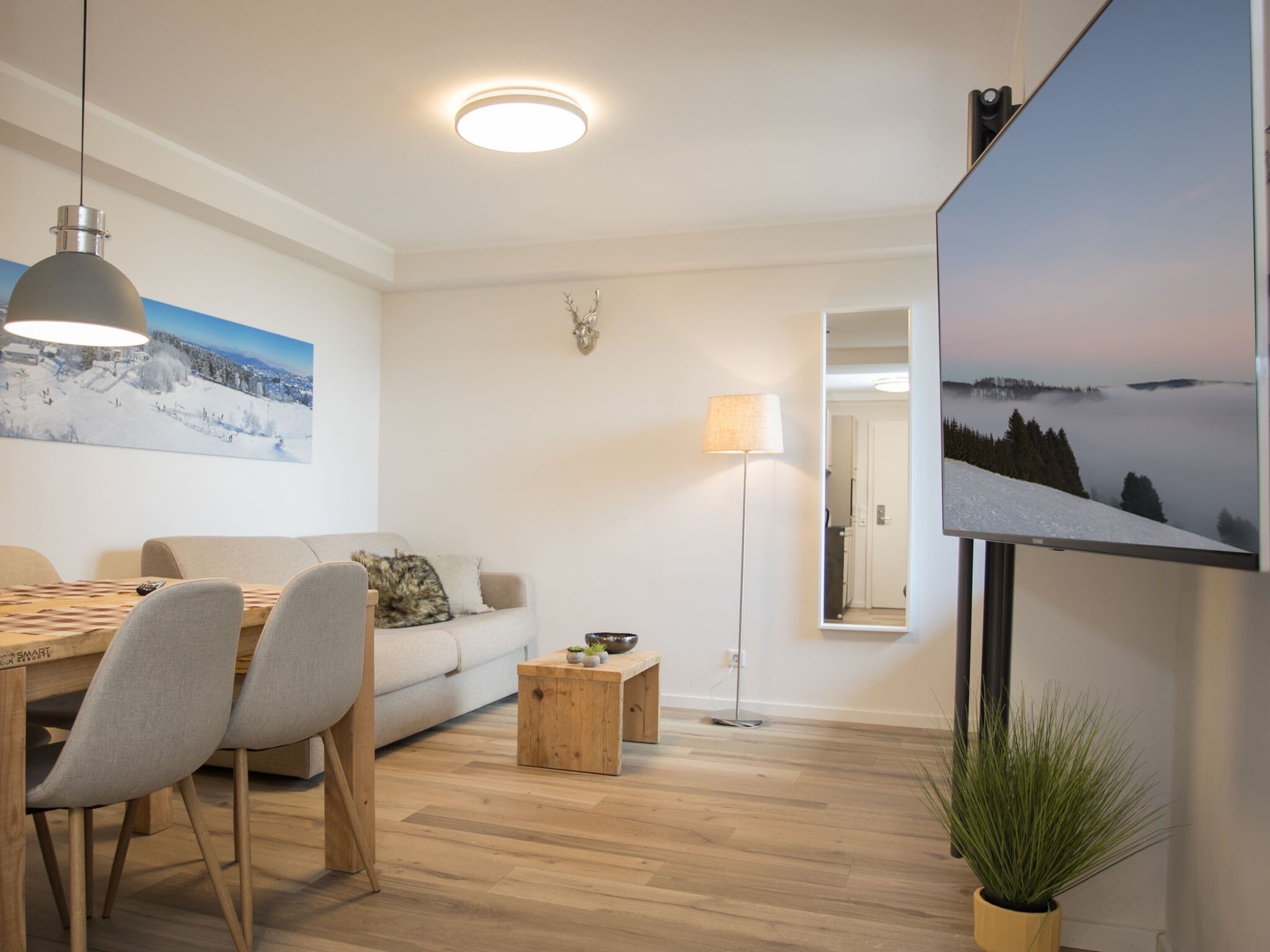 A modern living room with a wooden dining table, grey upholstered chairs, a sofa, a small wooden coffee table, a floor lamp, and a large flat-screen TV mounted on the wall.