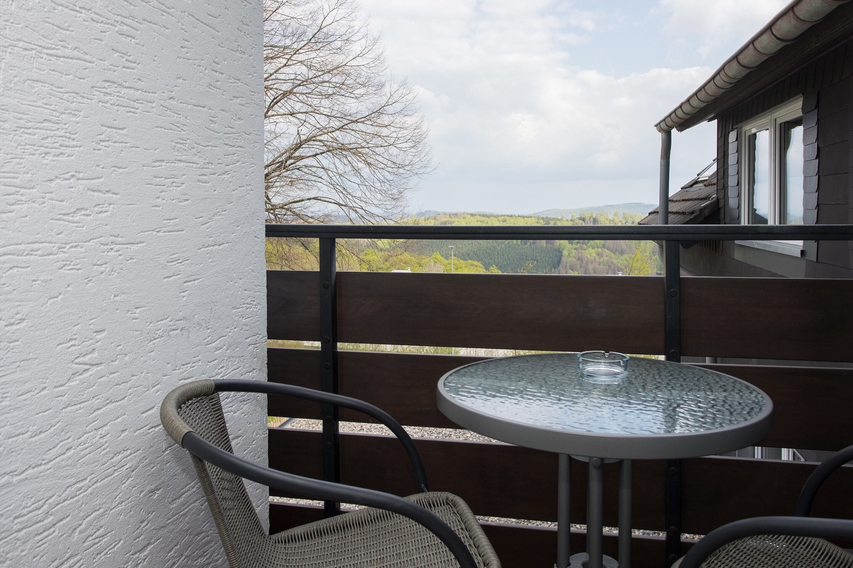 Un balcón con sillas de mimbre y una mesa de vidrio, con vistas a un paisaje boscoso.