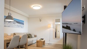 A modern living room with a wooden dining table, two chairs, a sofa, a floor lamp, and a flat-screen TV mounted on the wall. - Modern Flat in Neuastenberg With Balcony (Winterberg)