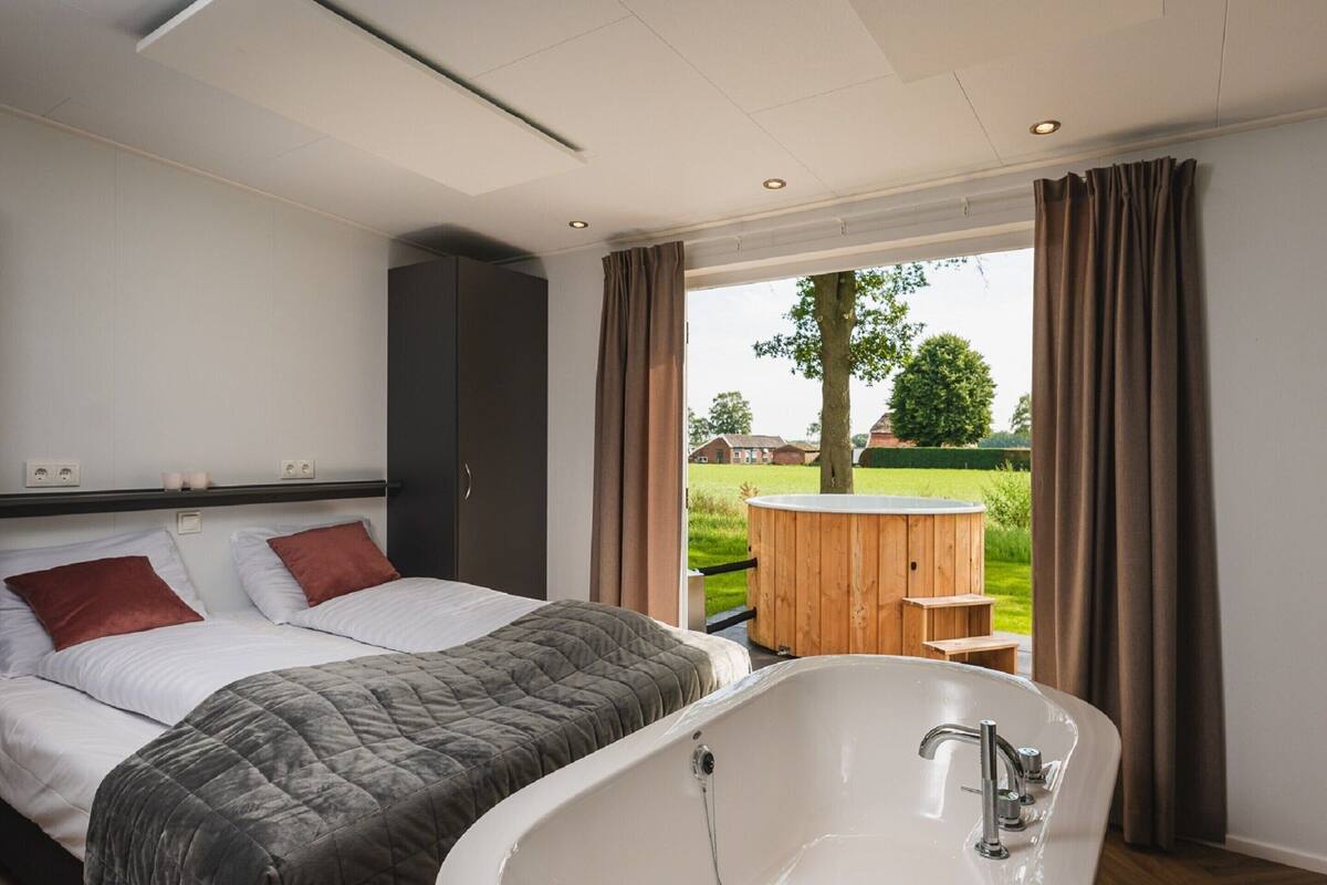 A bedroom with a large bed, a bathtub, and a view of a hot tub and garden.