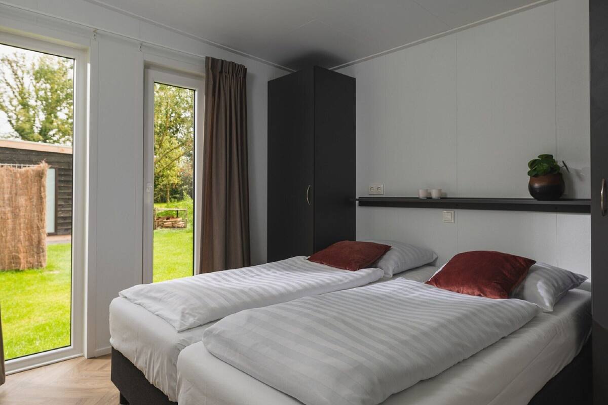 A bedroom with two beds, a dark wardrobe, a small shelf with cups, and a view of a garden through a sliding door.