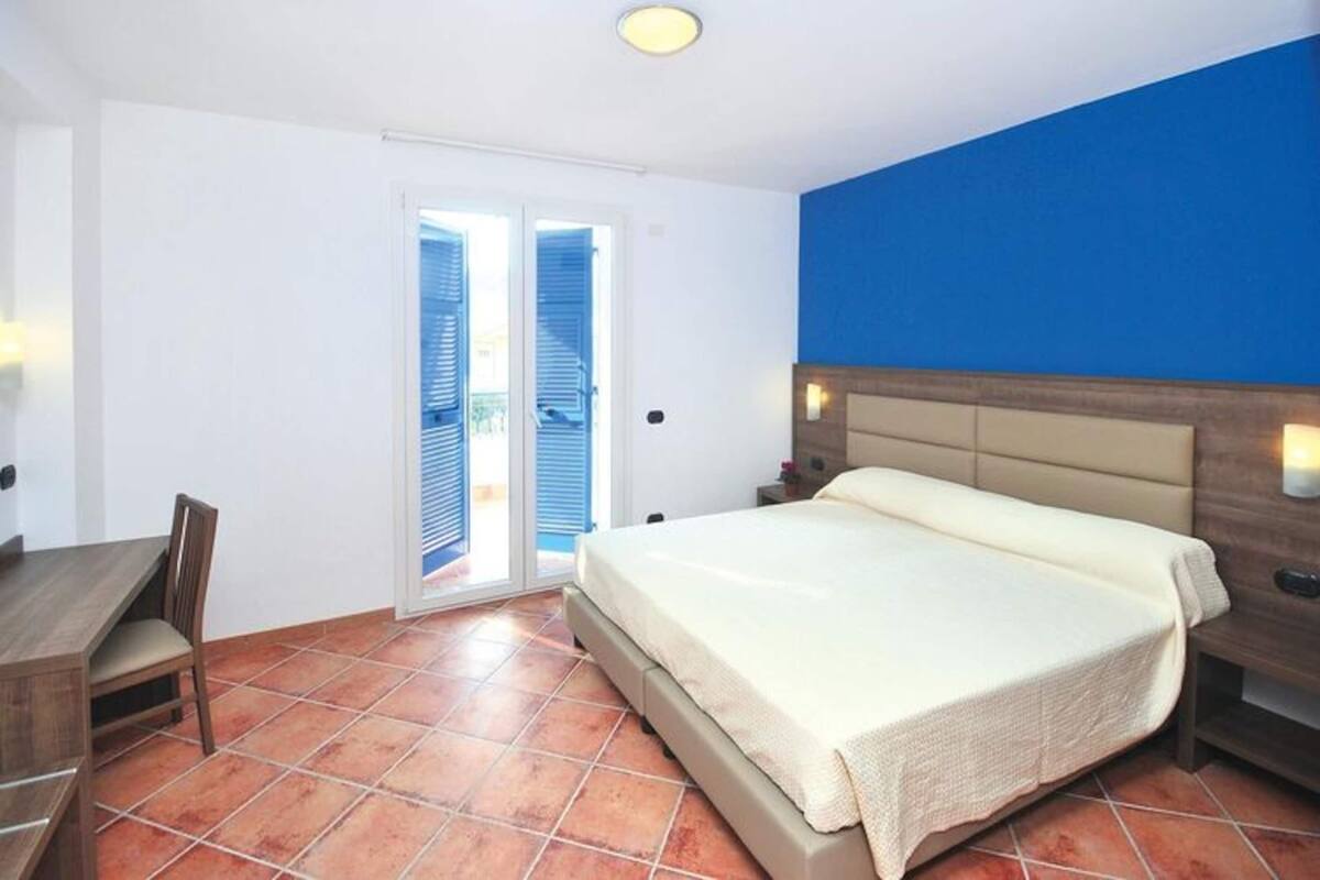 A bedroom with a bed, a desk, and a chair. There is a sliding door leading to a balcony.