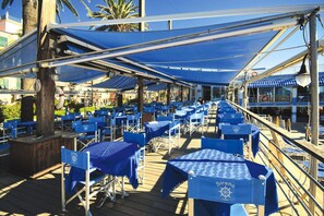 An outdoor dining area with blue tables and chairs, covered by blue awnings, and a view of the ocean and palm trees in the background.