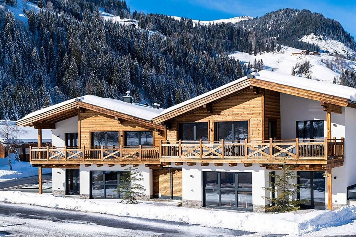 A two-story wooden chalet with balconies, large windows, and a snowy mountain backdrop.