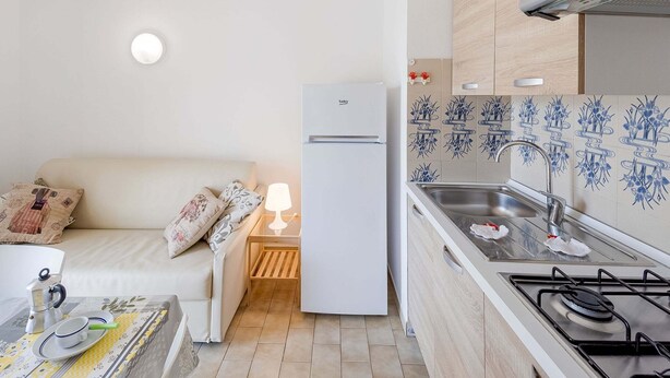 A compact kitchen with a refrigerator, stove, and sink. There's a small table with a lamp and a coffee maker. - Apartment in Loano With Garden (Loano)