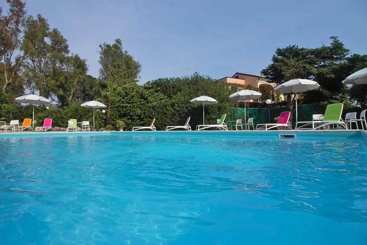 A swimming pool with lounge chairs and umbrellas, surrounded by trees and a building in the background.