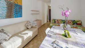 A modern living room with a beige sofa, a colorful sofa, a dining table with a floral tablecloth, and a large abstract painting on the wall. - Apartment in Loano With Garden (Loano)