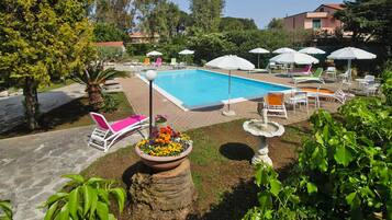 A swimming pool with lounge chairs and umbrellas, surrounded by greenery and a building in the background.