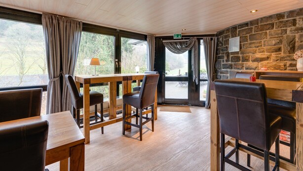 Un accogliente ristorante con tavoli e sedie in legno, una parete in pietra e ampie finestre che offrono vista sul panorama circostante.